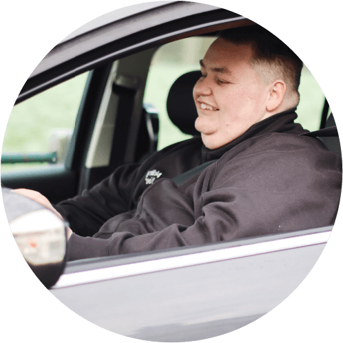 A person wearing a black jacket sits in the driver’s seat of a car, seen through the open window. They appear to be interacting with a device inside the vehicle. The seatbelt, dashboard, and part of the car interior are visible, with a blurred outdoor background.
